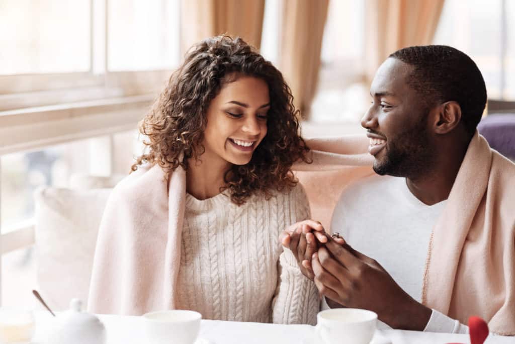 Smiling cheerful positive African American couple sitting in a cafe covered with a blanket while getting engaged.