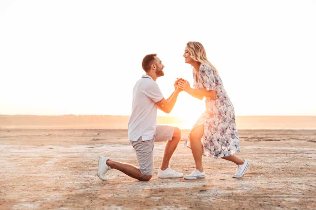 Photo of a happy man proposing to a woman with an engagement ring in a gift box while walking on a sunny beach.